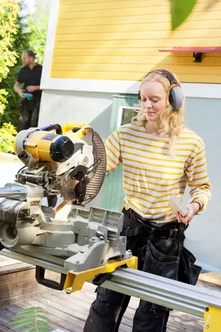 Tjej står och kapar en bräda vid ett gult hus. 